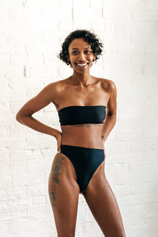 A woman standing with her hands on her hips smiling wearing high waisted minimal coverage black bikini bottoms with a matching black strapless bandeau bikini top.