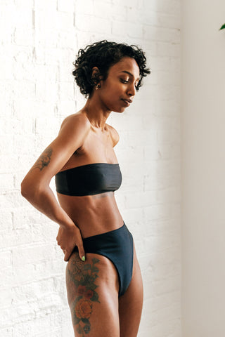 A woman standing to the side with her hands on her hips looking down wearing high waisted minimal coverage black bikini bottoms with a matching black strapless bandeau bikini top.