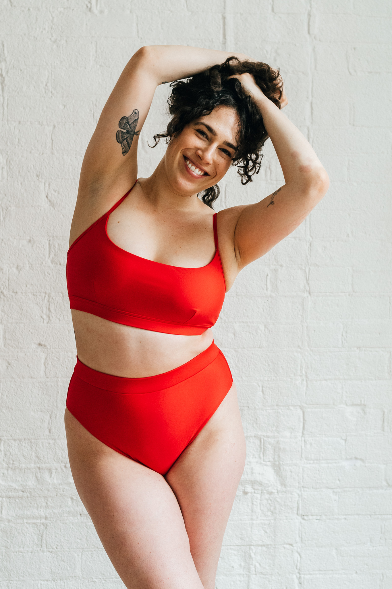 A woman leaning to the side with her hands in her hair wearing bright red high waisted bikini bottoms and a matching bright red bikini top with a scoop neckline.