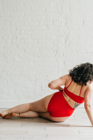 The back of a woman leaning against one arm on the ground wearing bright red high waisted bikini bottoms with full coverage and a matching bright red bikini top.