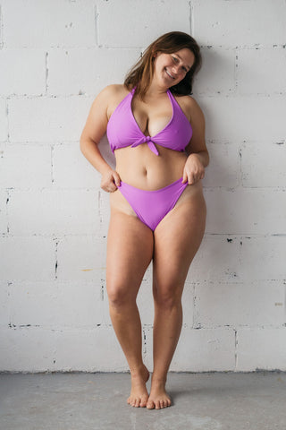 Woman smiling wearing a bright purple swimsuit with high cut bottoms and an adjustable tie front bikini top