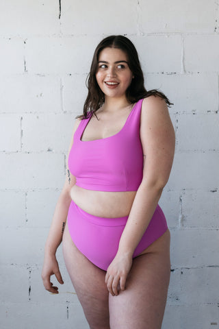 A woman standing in front of a white wall wearing a bright purple swimsuit featuring thick bikini straps for support and high waisted full coverage bottoms.