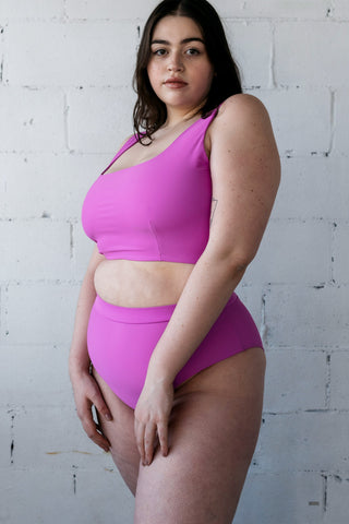 A woman standing in front of a white brick wall wearing bright orchid high waisted bikini bottoms with full coverage and a matching bright orchid bikini top.
