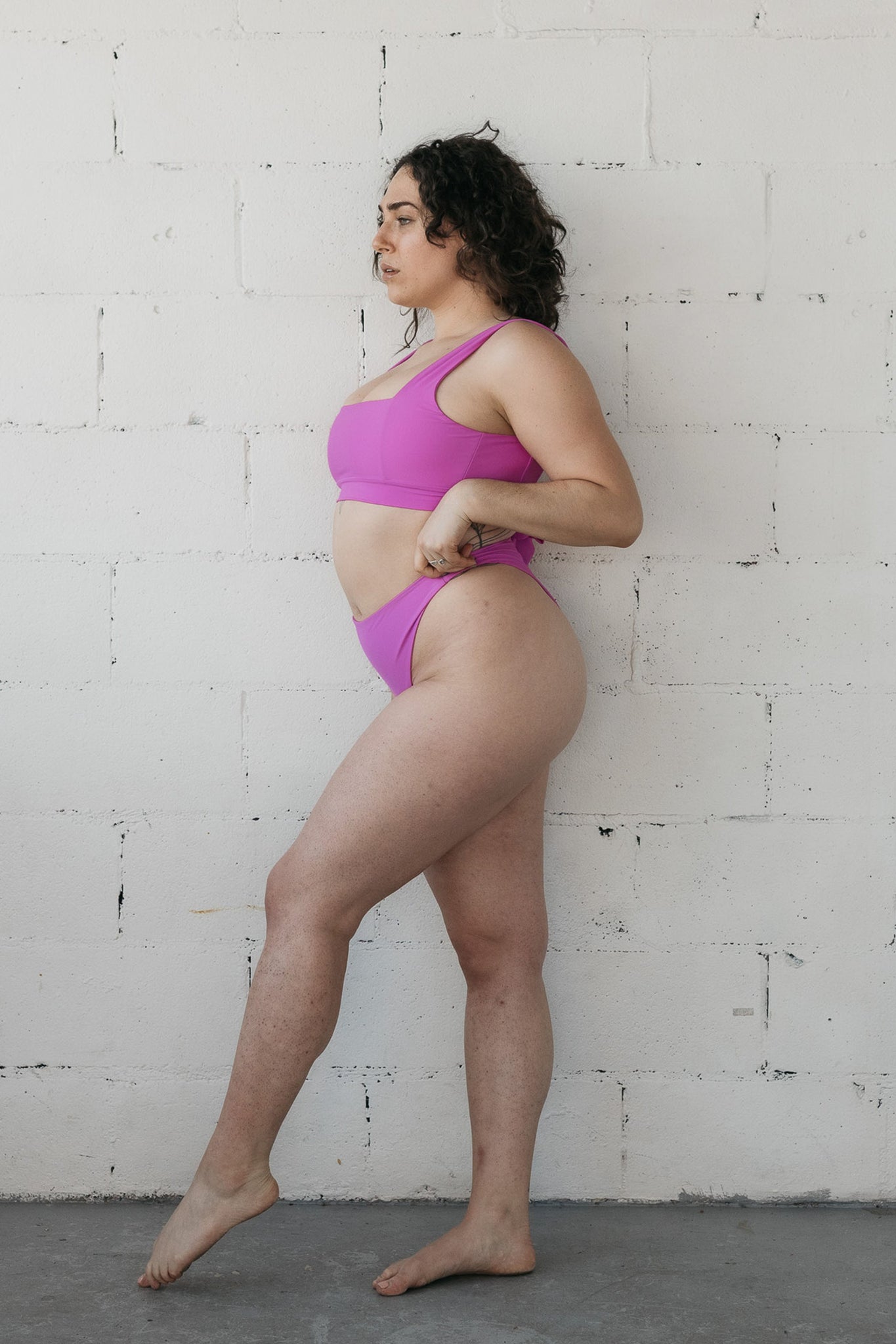 A woman standing to the side with one leg pointed in front of the other wearing bright pink high waisted bikini bottoms and a matching bright pink bikini top with a square neckline.