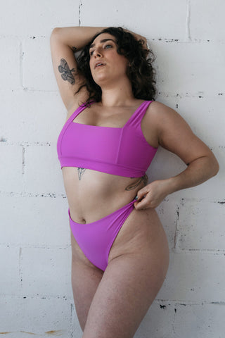 A woman leaning against a white brick wall with one hand in her hair wearing bright pink high waisted bikini bottoms and a matching bright pink bikini top with a square neckline.