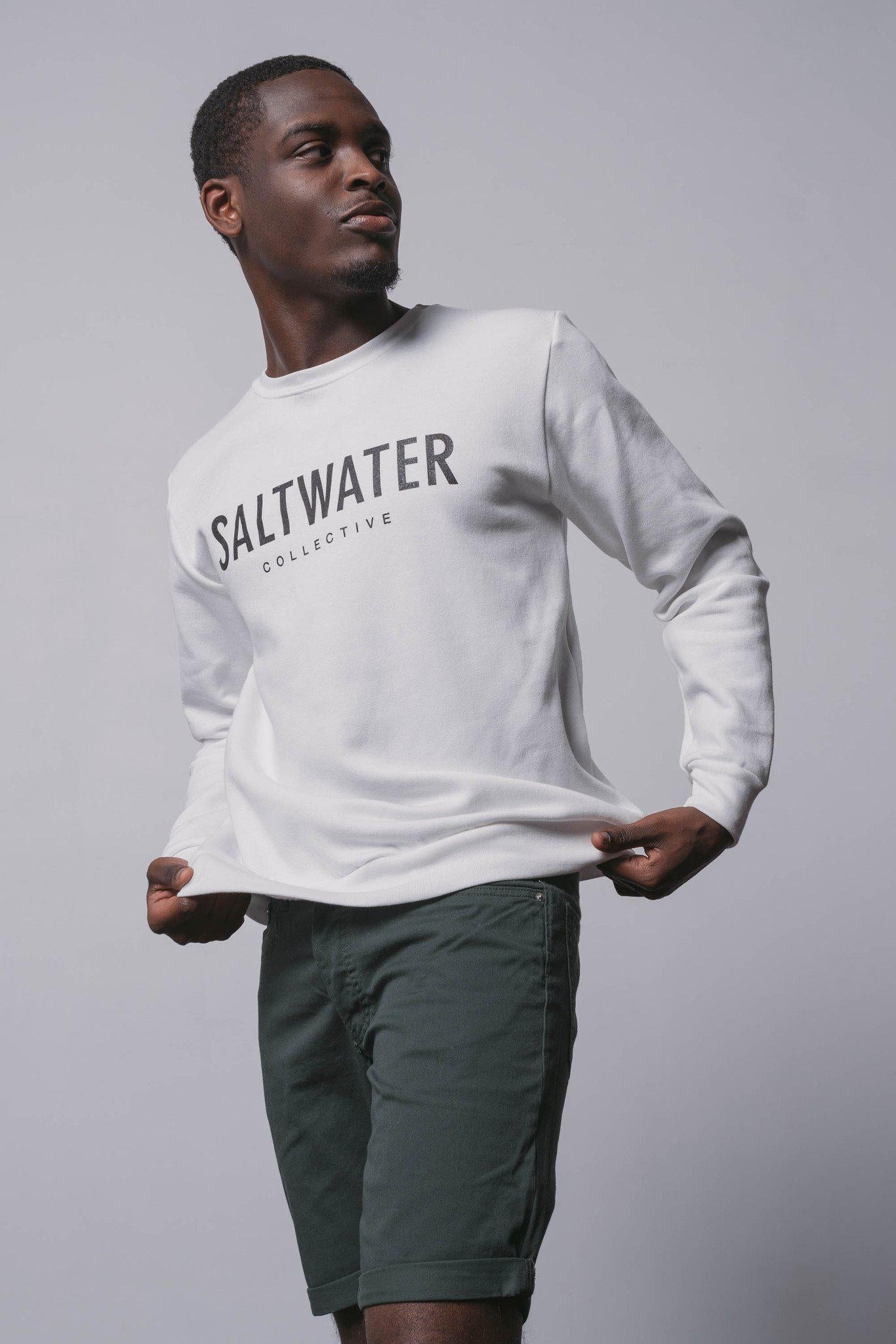 A guy standing in front of a white wall looking to the side and pulling a white crewneck sweatshirt down with a black SALTWATER logo printed across the chest.