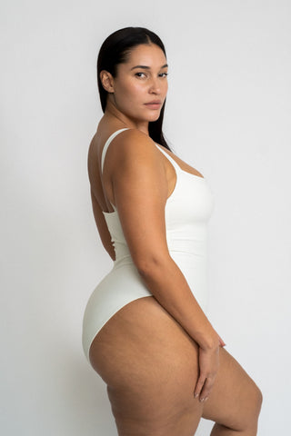 A woman standing to the side looking over her shoulder wearing a white one piece swimsuit with a straight neckline.