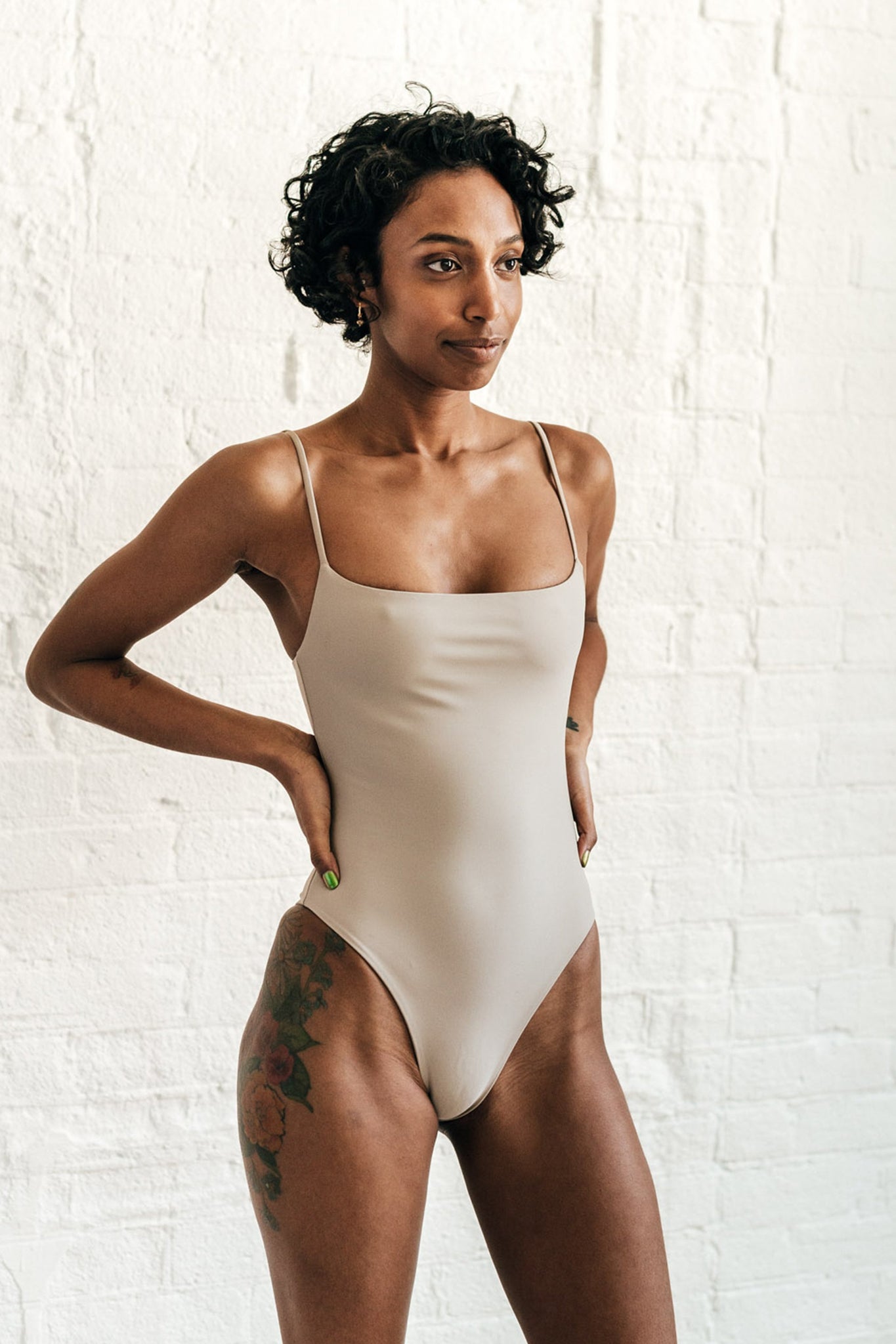 A woman standing with her hands on her hips wearing a nude one piece swimsuit with spaghetti straps.