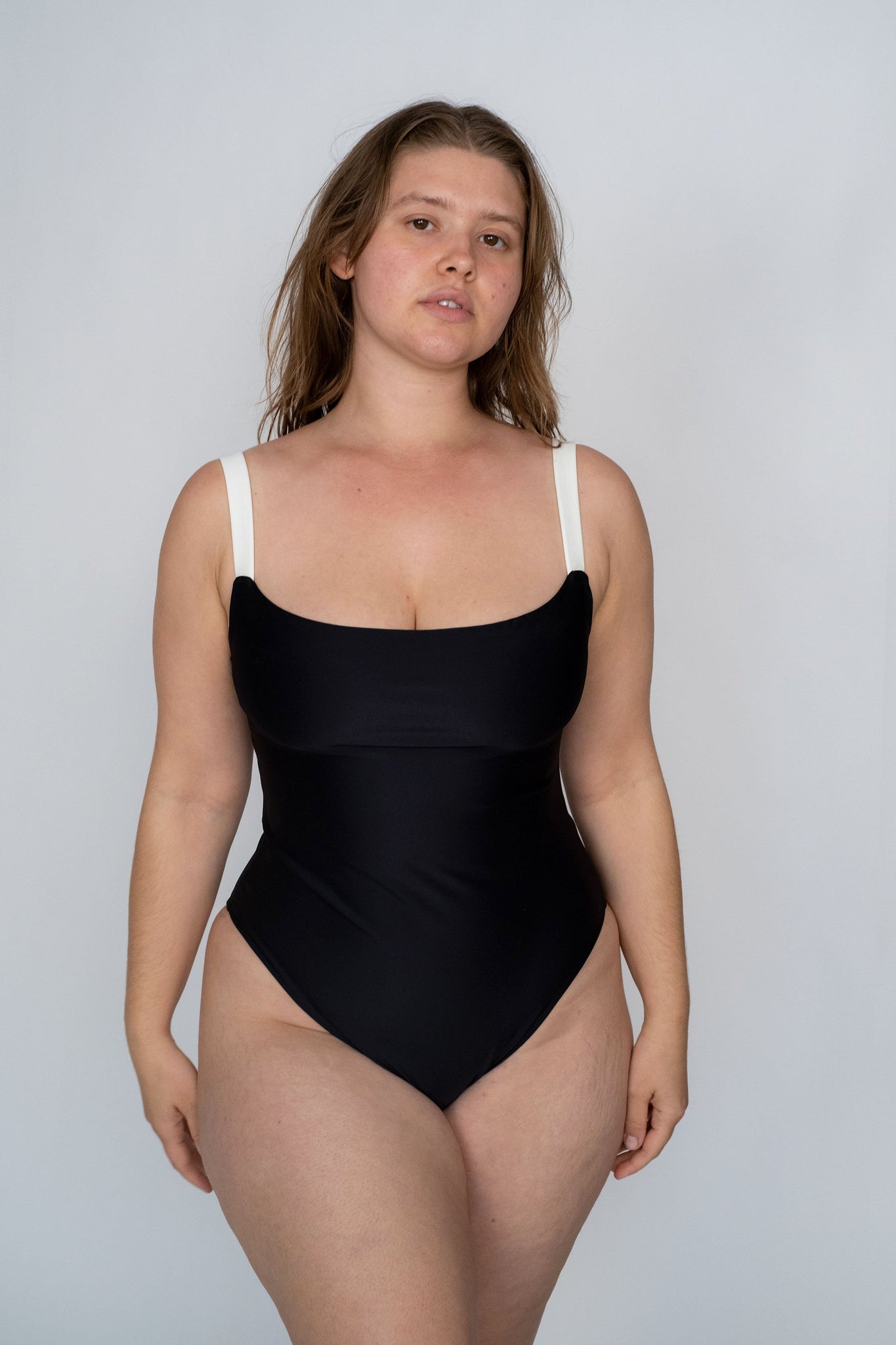 A woman standing wearing a black one piece swimsuit with white thick spaghetti straps.
