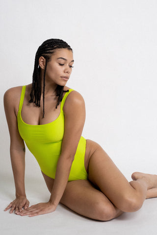 A woman sitting on the ground leaning forward wearing a neon green one piece swimsuit with a straight neckline.