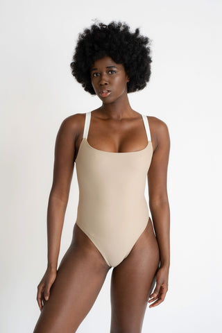 A woman standing wearing a nude one piece swimsuit with thick white spaghetti straps.