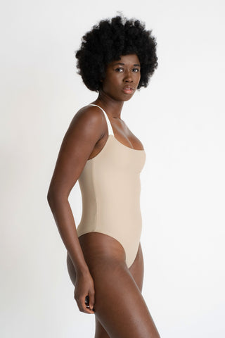 A woman standing to the side wearing a nude one piece swimsuit with thick white spaghetti straps.