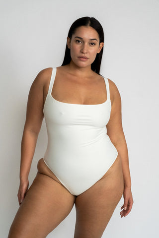 A woman standing with one leg pushed to the side wearing a white one piece swimsuit with a straight neckline.