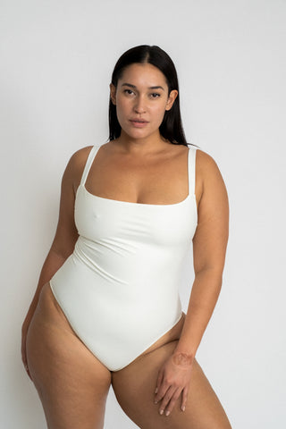A woman standing leaning on one hip wearing a white one piece swimsuit with a straight neckline.