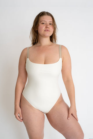 A woman standing with one leg bent wearing a white one piece with thick nude spaghetti straps.