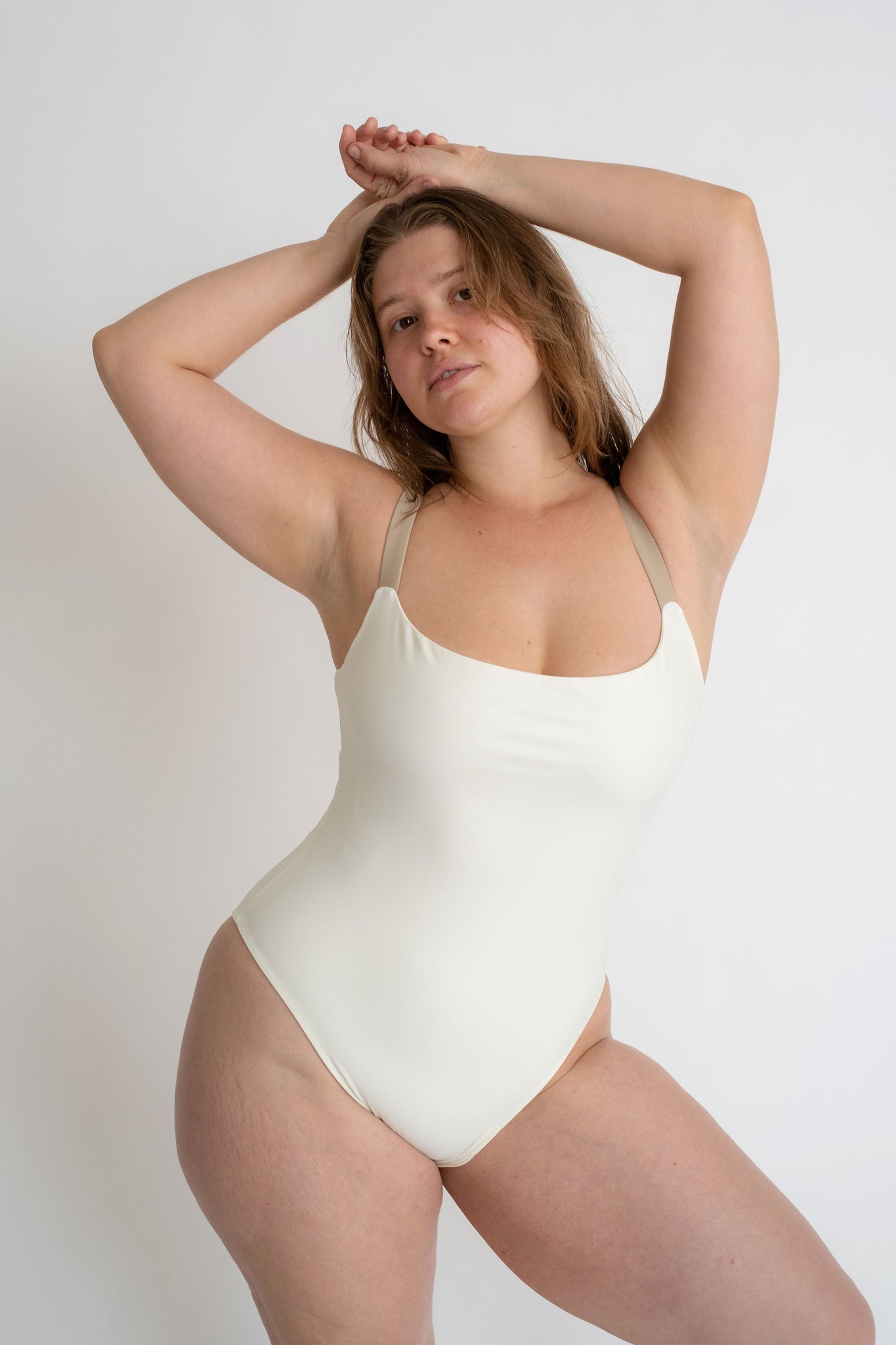 A woman standing with her hands above her head wearing a white one piece with thick nude spaghetti straps.