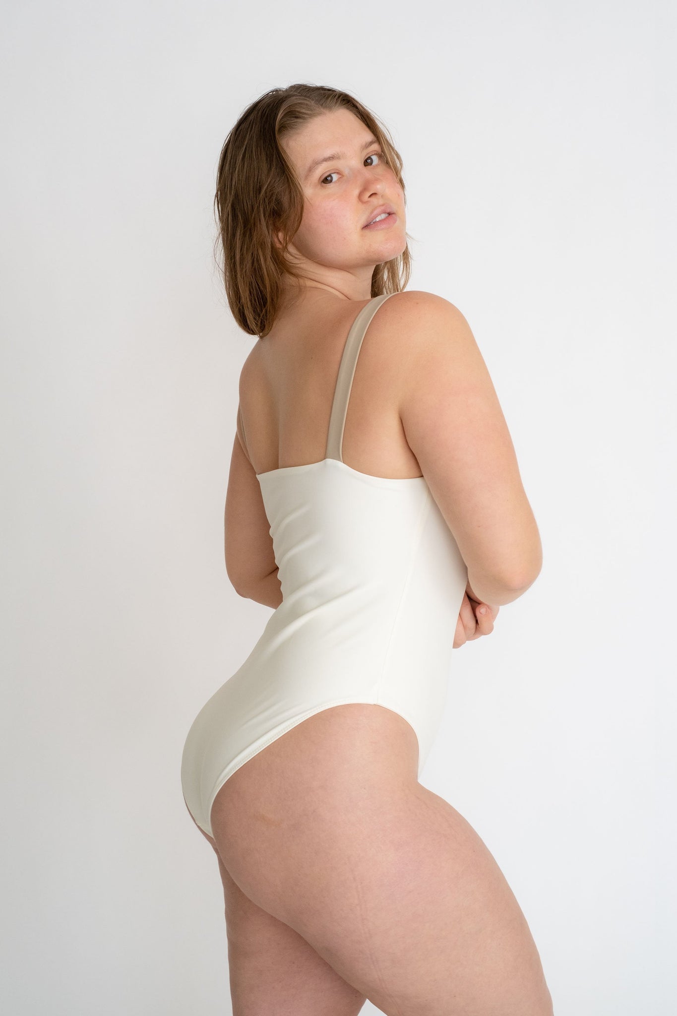 A woman standing to the side looking over her shoulder wearing a white one piece with thick nude spaghetti straps.