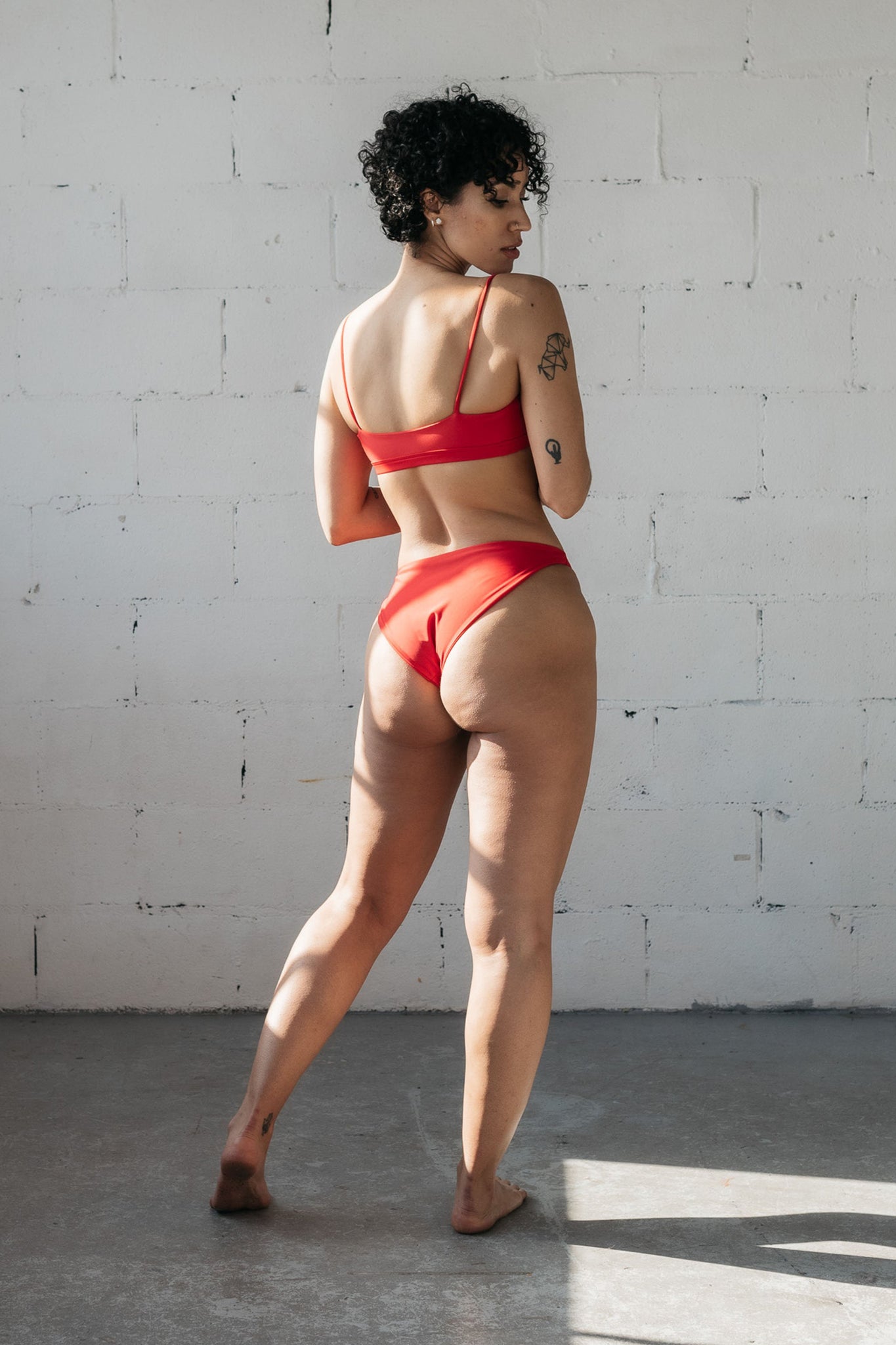 A woman standing and looking over her shoulder wearing high cut red bikini bottoms and a red bikini top with spaghetti straps.