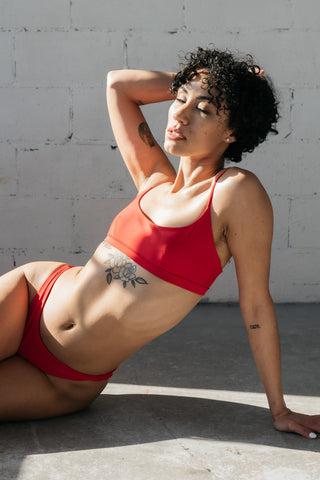 A woman leaning on her arm on the floor and one arm in her hair wearing a red swimsuit with high cut bikini bottoms and a scoop neck bikini top. 