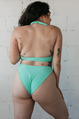 The back of a woman standing in front of a white wall wearing turquoise high cut bikini bottoms with a matching turquoise bikini top.