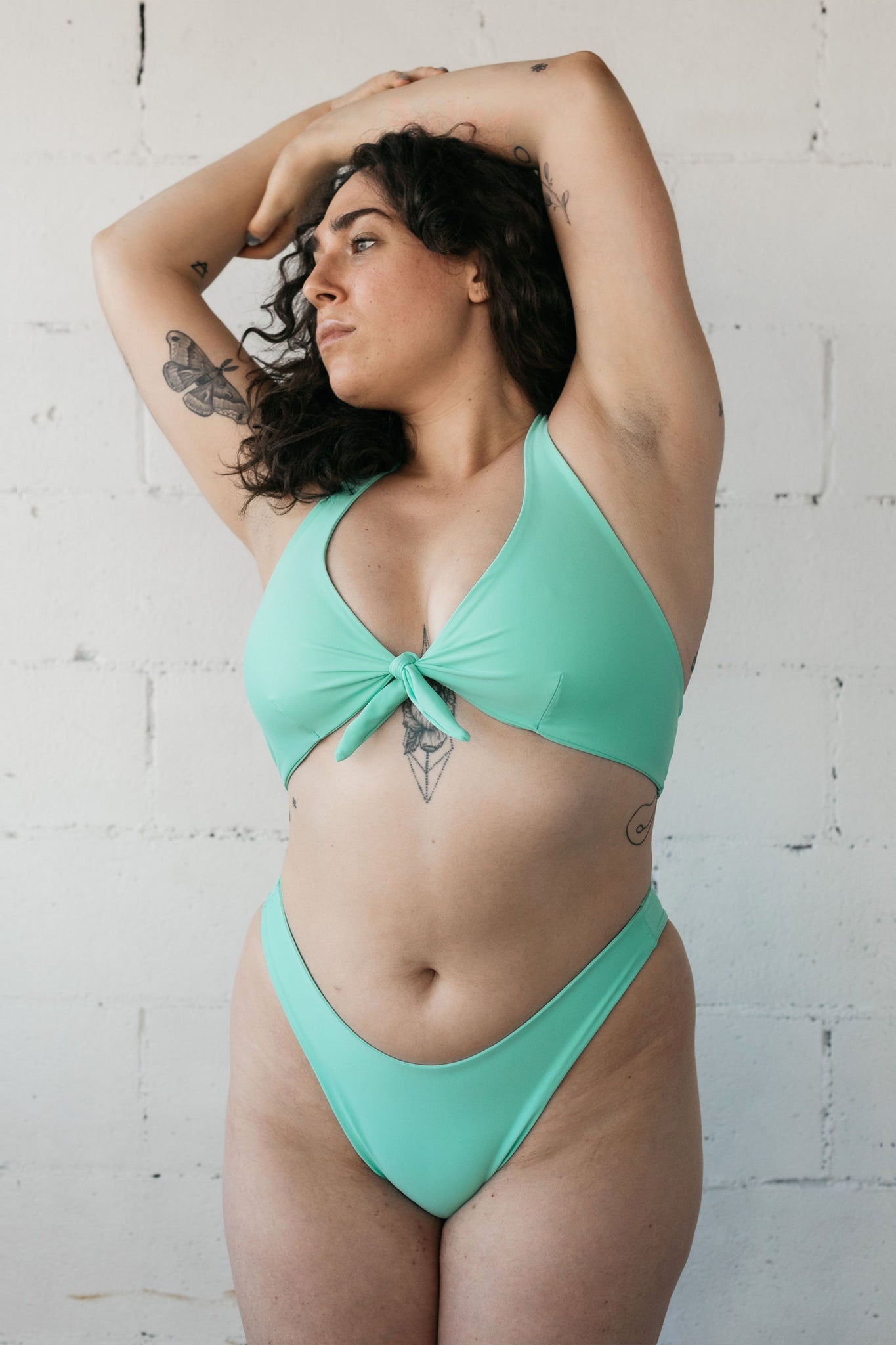 A woman standing with her arms crossed above her head wearing high cut turquoise bikini bottoms and a matching tie front turquoise bikini top.