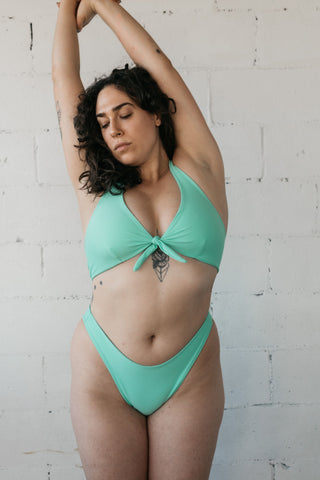 Woman stretching arms above her head wearing a seafoam turquoise swimsuit with high cut bikini bottoms and adjustable front bikini top