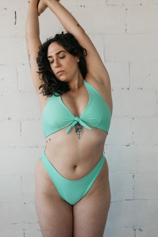 A woman leaning to the side with her arms stretched above her wearing high cut turquoise bikini bottoms and a matching tie front turquoise bikini top.