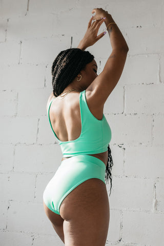 A woman standing in front of a white brick wall with her hands above her head wearing turquoise high waisted full coverage bikini bottoms with a matching turquoise bikini top.