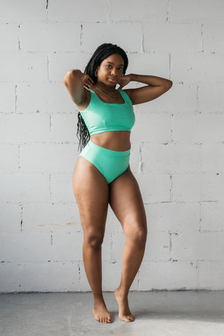 A woman standing with her hands resting on her shoulders wearing turquoise high waisted full coverage bikini bottoms with a matching turquoise bikini top.