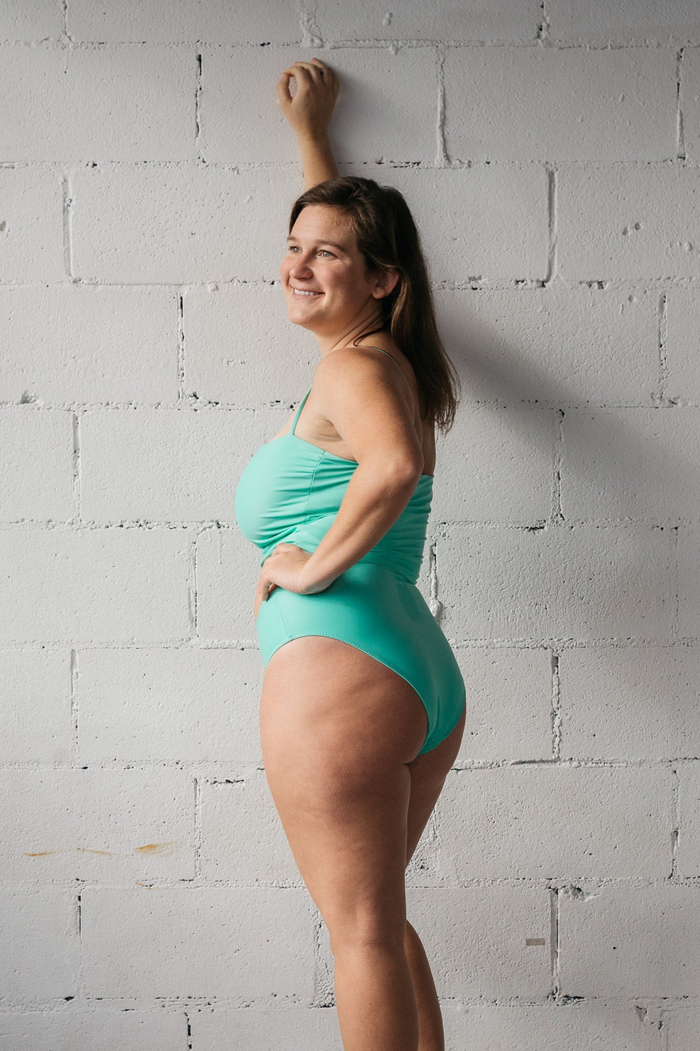 A woman standing with one hand against a white wall wearing turquoise high waisted bikini bottoms and a matching turquoise tankini top.