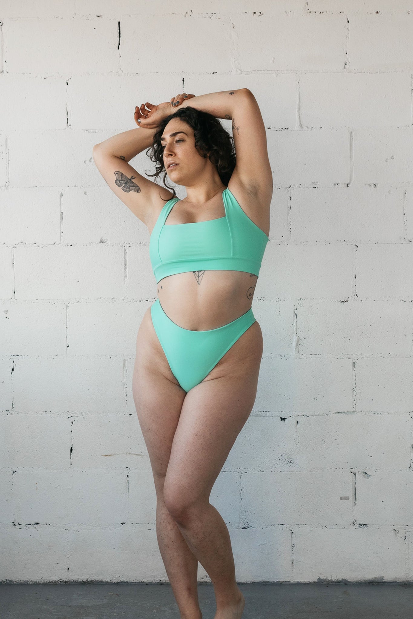 A woman standing with her hands crossed above her head wearing high cut turquoise bikini bottoms and a matching turquoise bikini top with a square neckline.