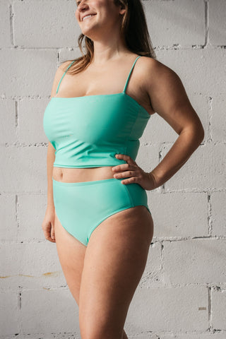 A woman standing with one hand on her hip wearing turquoise high-waisted bikini bottoms with a matching turquoise tankini top.