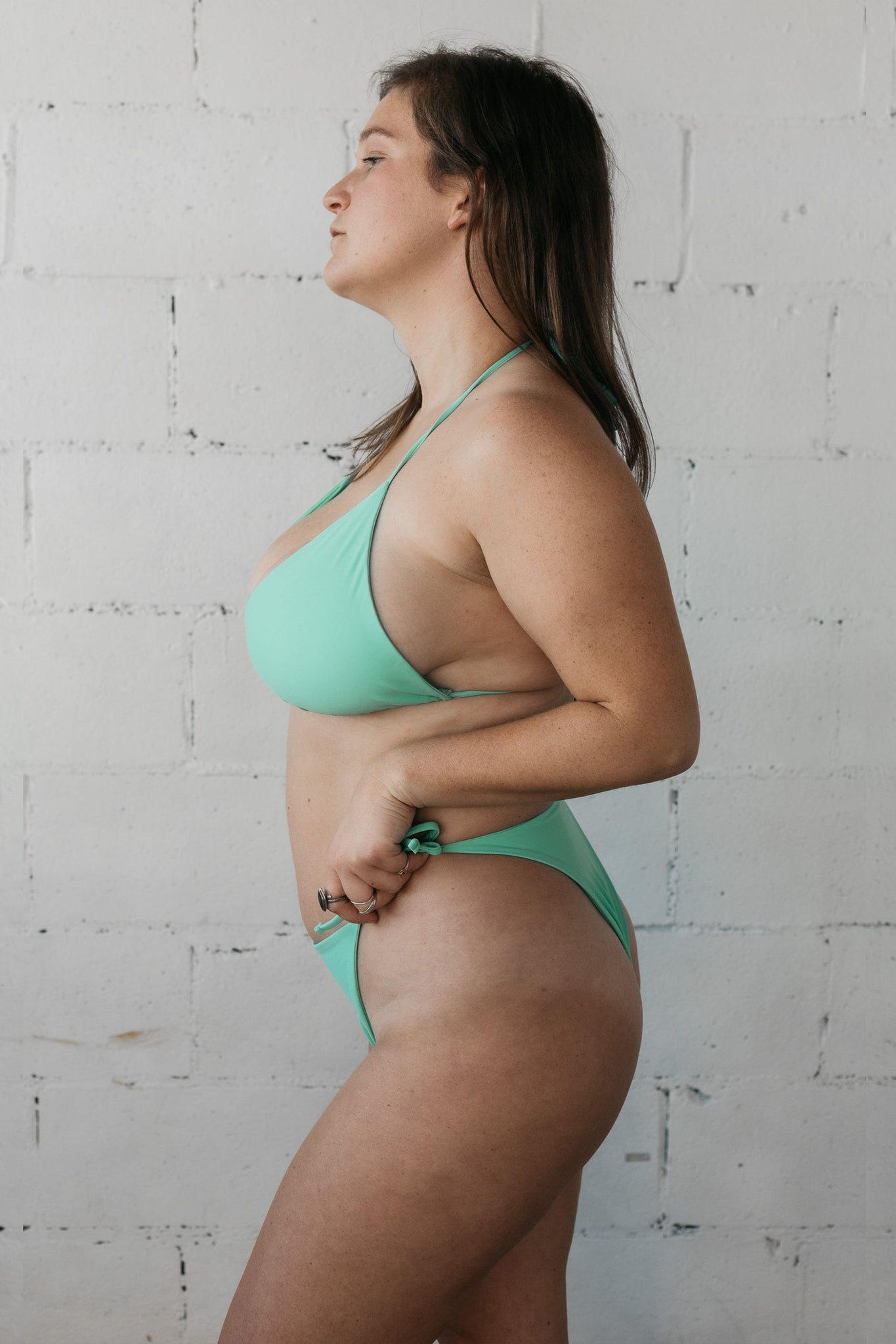 A woman standing to the side with her hands on her hips wearing turquoise triangle string bikini bottoms with a matching turquoise adjustable triangle string bikini top.