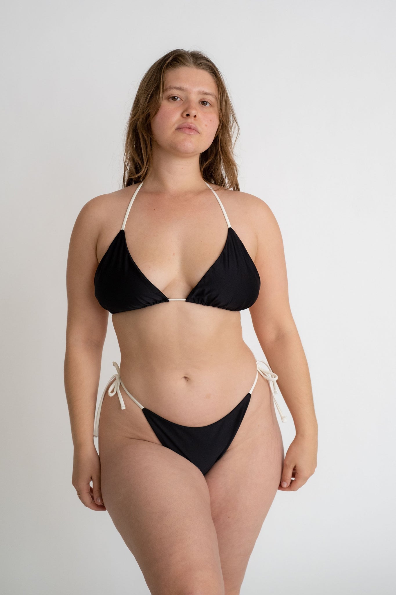 A woman standing with her arms relaxed by her sides wearing a black triangle bikini top with adjustable white straps and black triangle bikini bottoms with white strings.