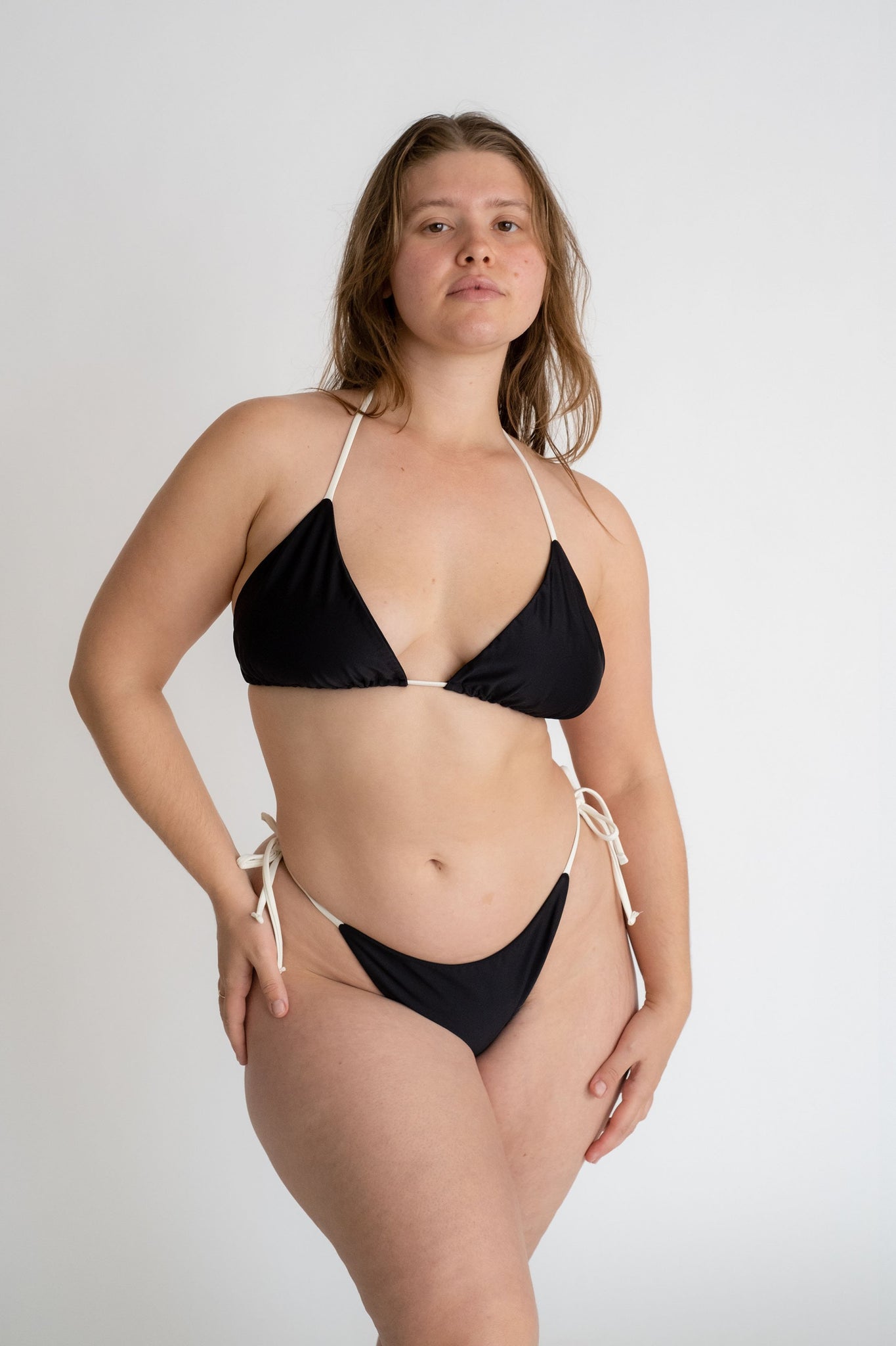 A woman standing with one hand on her hip wearing black triangle bikini bottoms with white string ties and a matching black triangle bikini top with adjustable white strings.