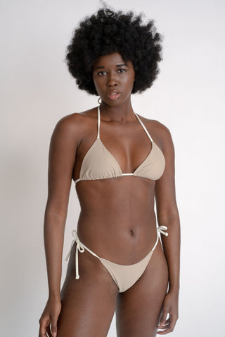 A woman standing with her arms relaxed by her sides wearing nude bikini bottoms with white adjustable strings and a matching nude triangle bikini top with white adjustable strings.