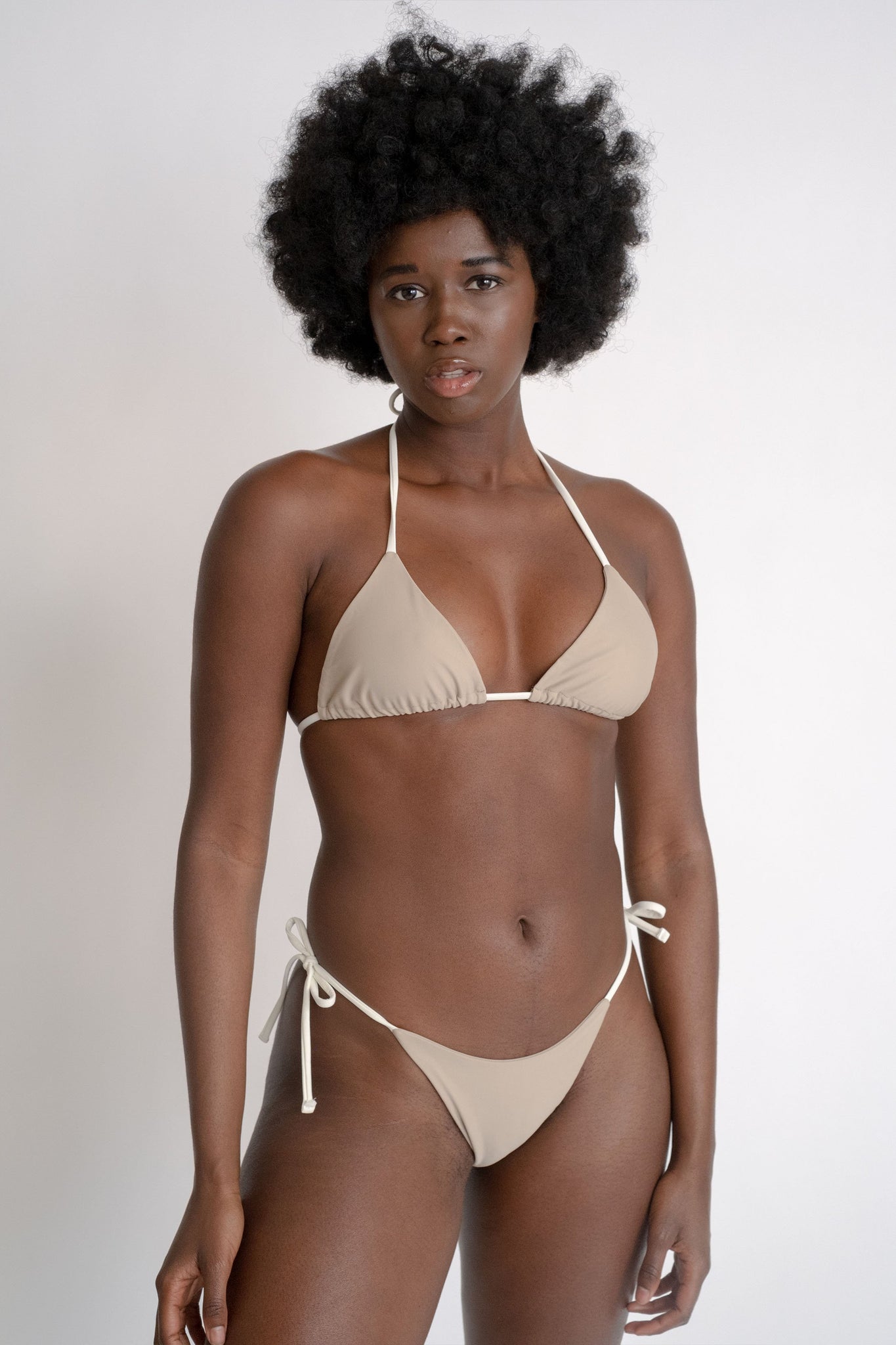 A woman standing with her arms relaxed by her sides wearing nude triangle bikini bottoms with adjustable white strings and a matching nude triangle bikini top with white adjustable strings.