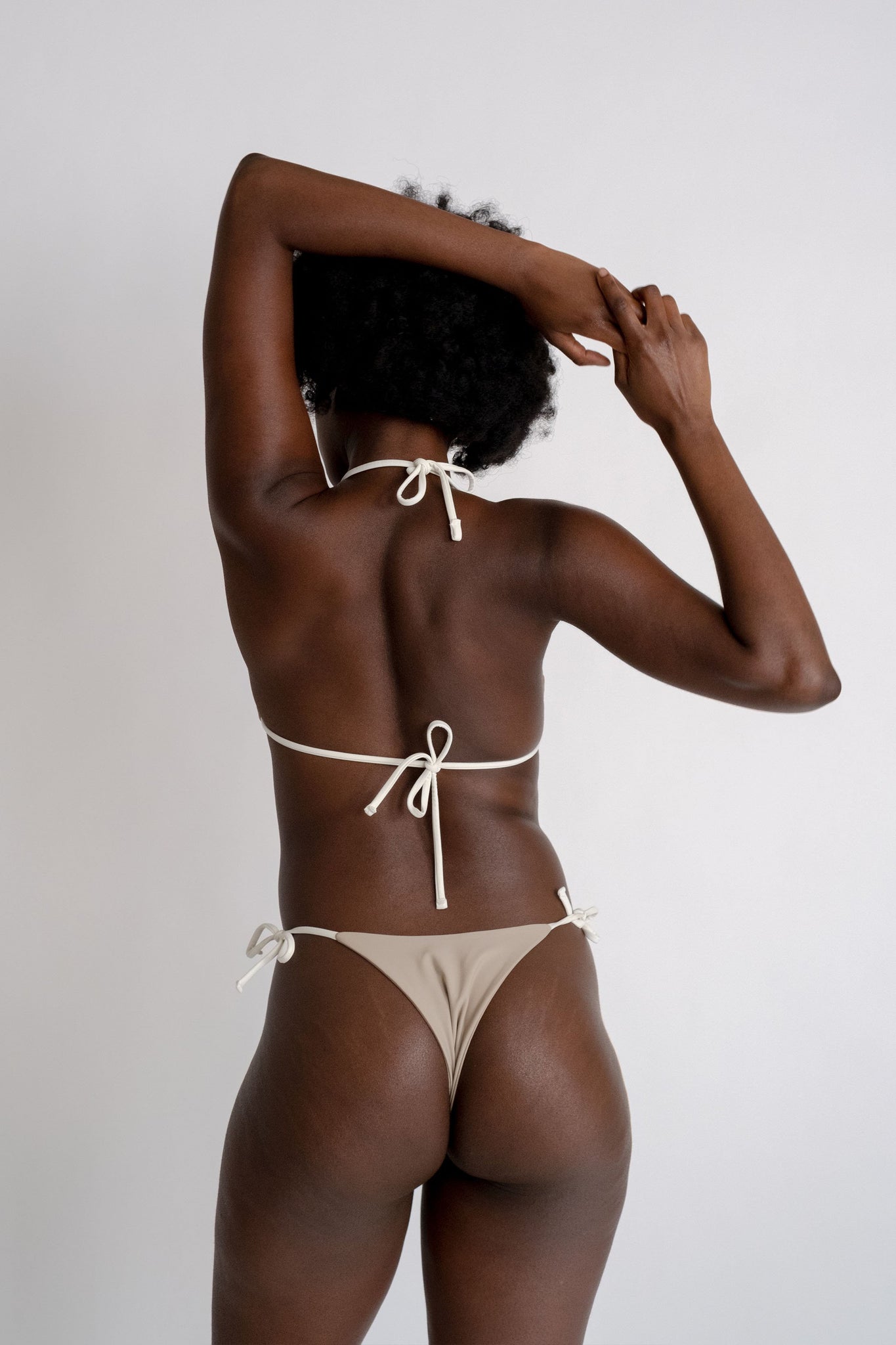 The back of a woman with her arms folded wearing nude bikini bottoms with white adjustable strings and a matching nude triangle bikini top with white adjustable strings.