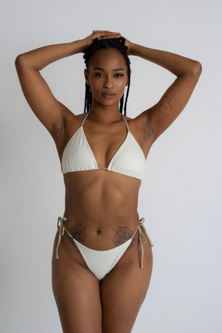 A woman standing with her hands folded above her head wearing white triangle bikini bottoms with nude adjustable strings with a matching white triangle bikini top and nude adjustable straps.