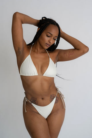 A woman standing with her arms folded on her head looking down wearing white triangle bikini bottoms with nude adjustable strings with a matching white triangle bikini top and nude adjustable straps.