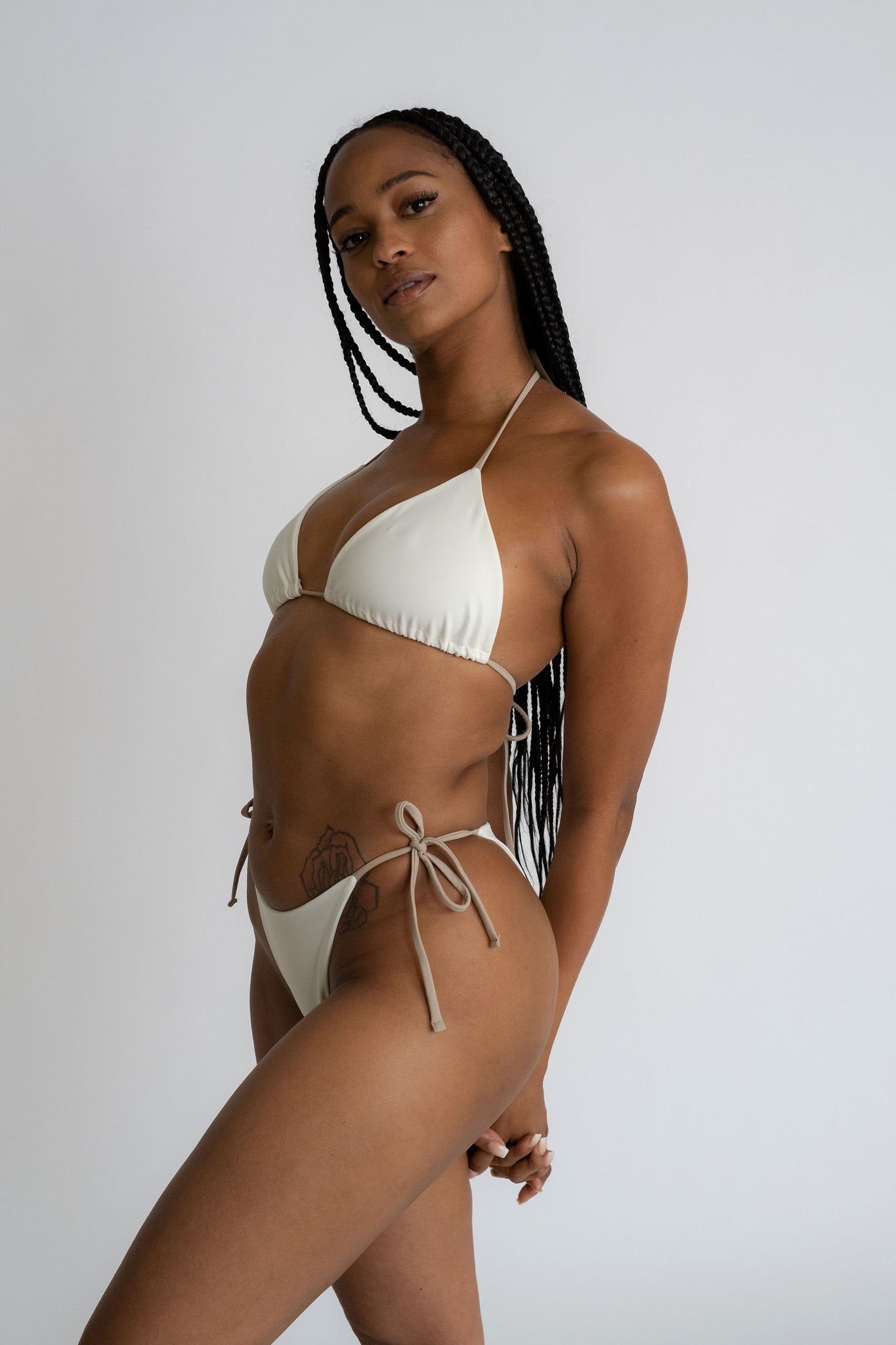 A woman standing with her arms wrapped behind her wearing white triangle bikini bottoms with adjustable nude string ties and a matching white bikini top with nude string ties.