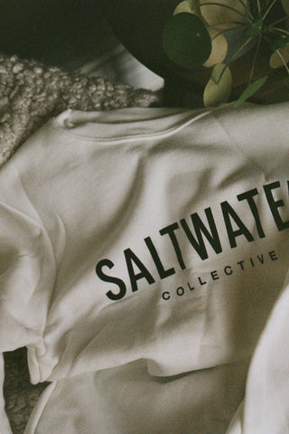 A close up of a white crewneck sweatshirt with a SALTWATER logo printed across the chest in black.